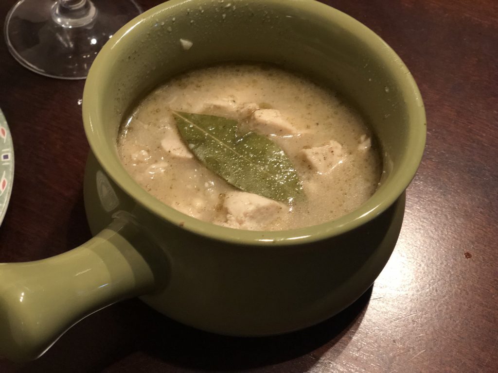 A hearty bowl of chicken and barley stew served in a green ceramic soup mug, topped with a bay leaf. The warm soup sits on a dark wooden table, ready for a comforting meal.