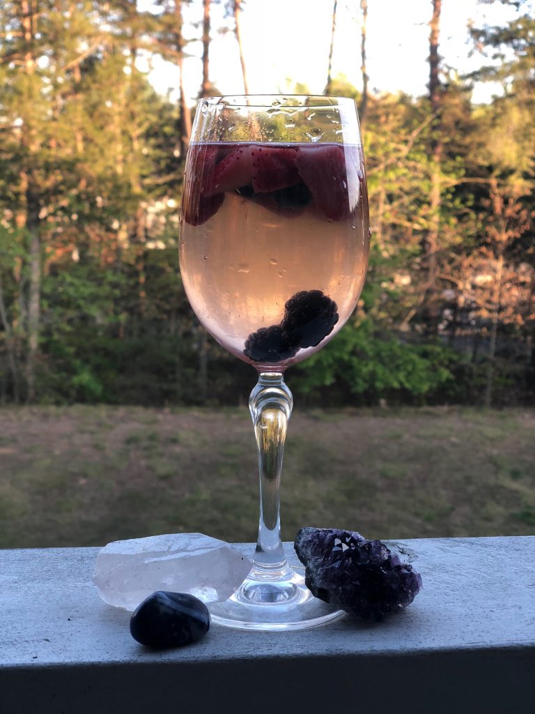 A glass of Beltane berry sangria filled with fresh strawberries, blueberries, and blackberries, placed outdoors on a ledge with a backdrop of spring greenery. Crystals, including amethyst, obsidian, and clear quartz, are arranged at the base of the glass, adding a magical touch.