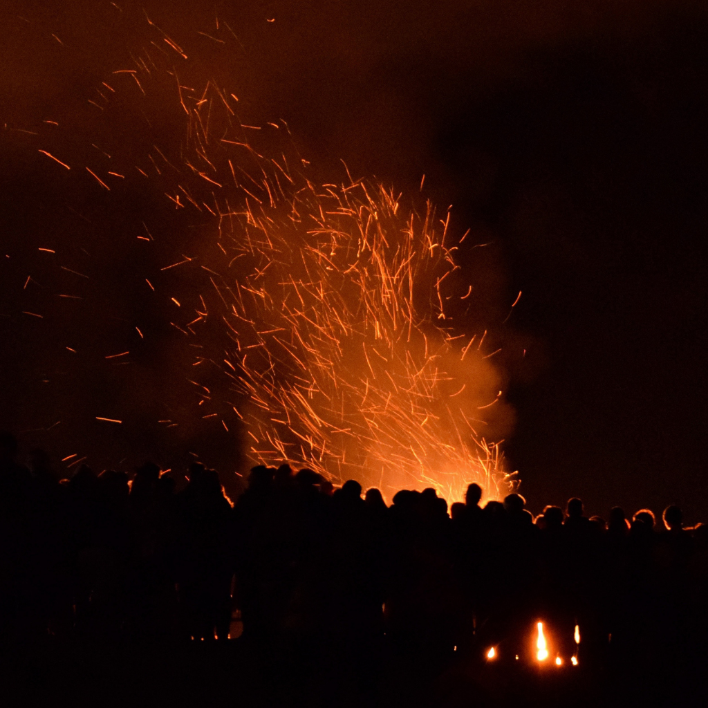 A large Beltane bonfire at night, surrounded by a silhouetted crowd. Sparks and embers rise into the dark sky, highlighting the fire's warmth and energy.