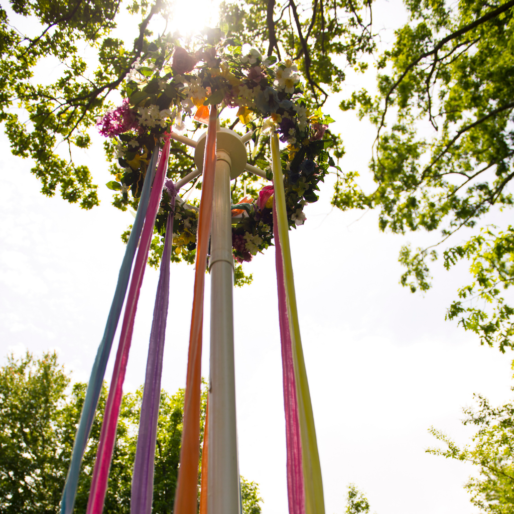 A traditional Beltane Maypole adorned with a floral wreath at the top and colorful ribbons flowing down, set against a bright, sunny sky framed by green leafy trees.