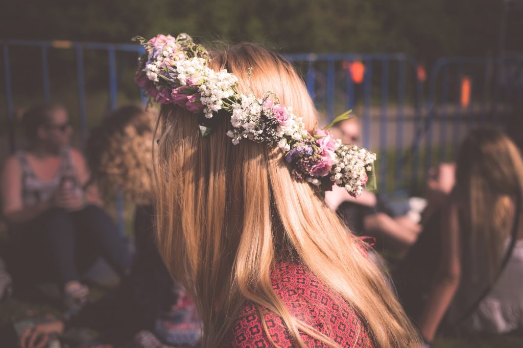 A blonde woman wearing a floral crown with pink roses and white baby's breath sits outdoors among a group of people at a springtime gathering. The warm sunlight highlights her hair, creating a festive and serene atmosphere.