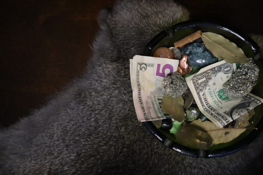 A money bowl filled with cash, pyrite, bay leaves, and crystals resting on a soft fur backdrop, designed to attract abundance and prosperity through custom spellwork.