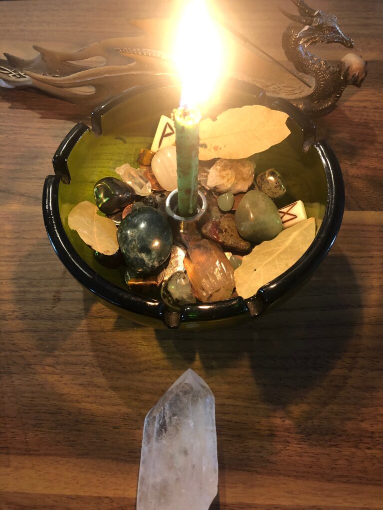 A money bowl featuring a green candle burning brightly at the center, surrounded by crystals, coins, and bay leaves, all placed inside a green dish. A quartz crystal point sits at the forefront, adding a touch of intention and abundance.