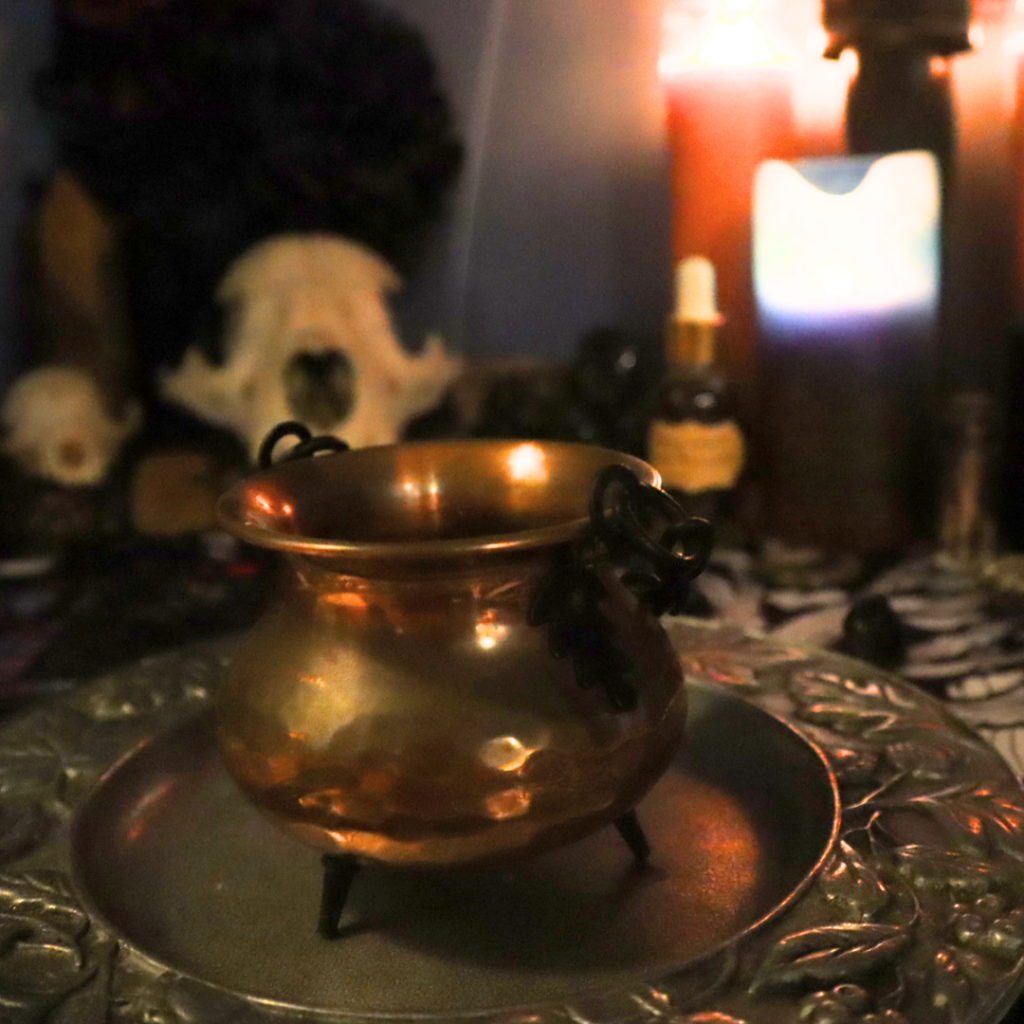 A glowing copper cauldron sits on an ornate silver tray, surrounded by a mystical altar featuring candles, skull decor, and magical tools. The warm lighting creates a magical and personalized ambiance.