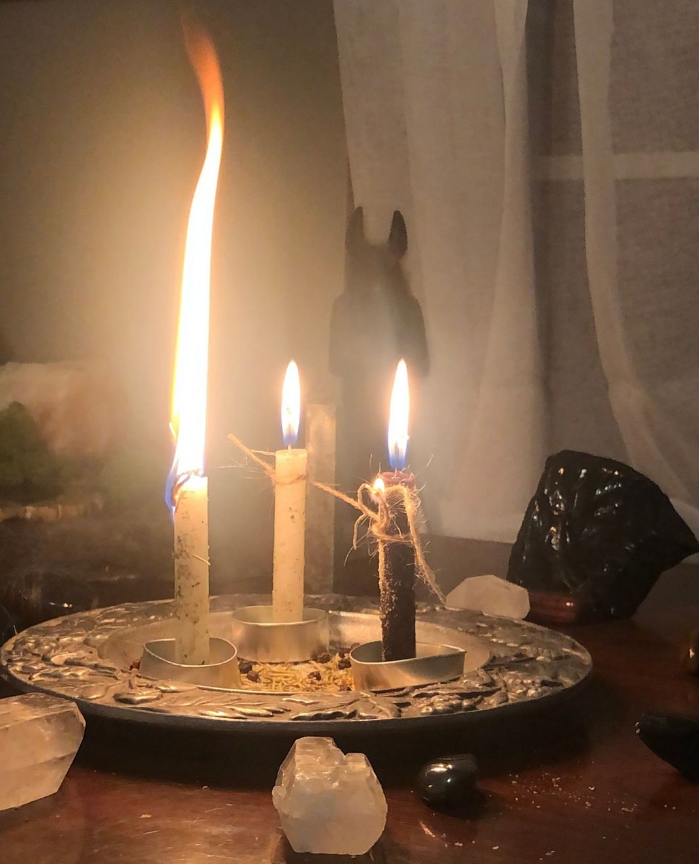 A ritual setup featuring three burning candles—one with a tall flame, tied with twine—arranged on a silver plate with herbs, crystals, and a dark gemstone on a wooden table. A tall dark statue of Anubis looms in the background. The warm, mystical ambiance evokes custom spellwork tailored to individual needs.